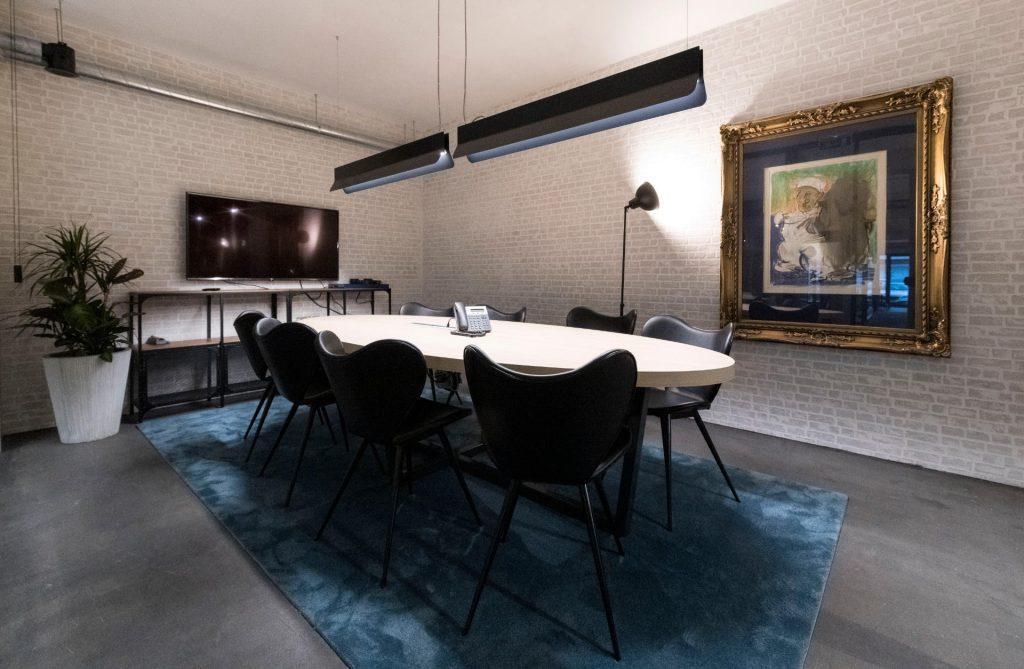 Vista de outra sala de reuniões no Outglocal Coworking Aveiro, com mesa oval, tapete azul e parede de tijolo branco.