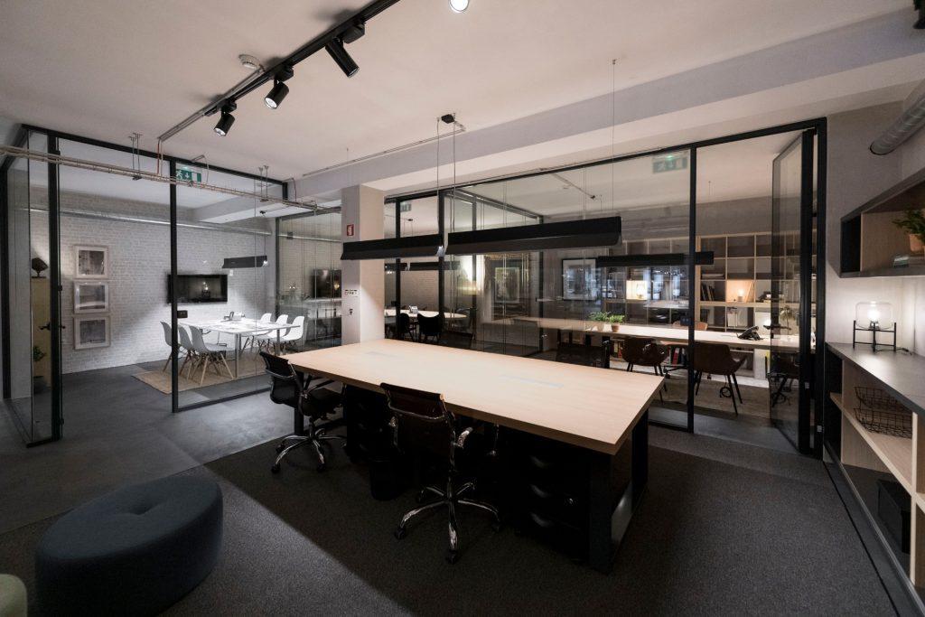 Vista de uma mesa de trabalho em open space no Outglocal Coworking Aveiro, com gabinetes privados com divisórias em vidro ao fundo.